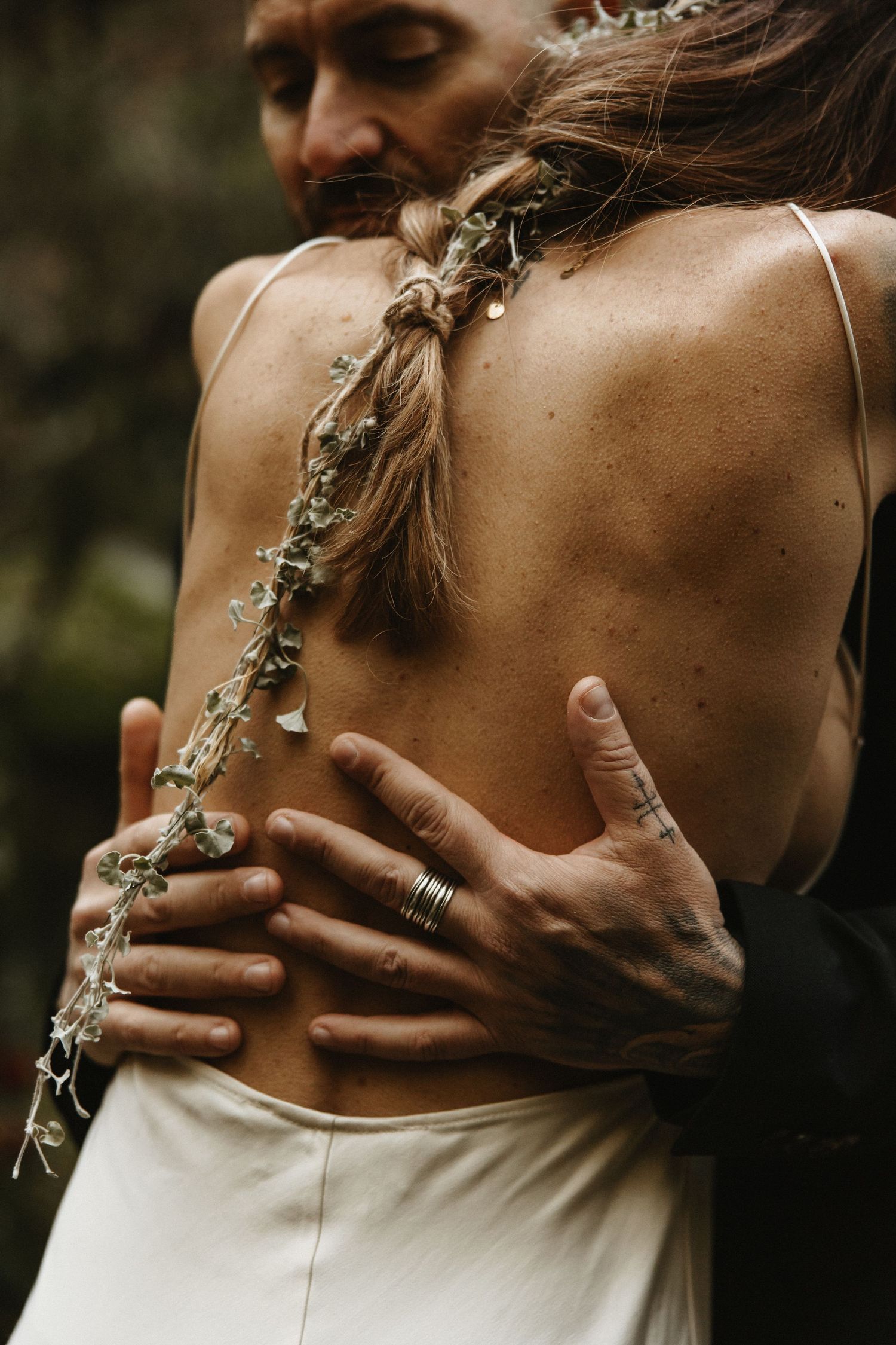 Groom embracing bride in backless wedding dress with floral braid in forest.