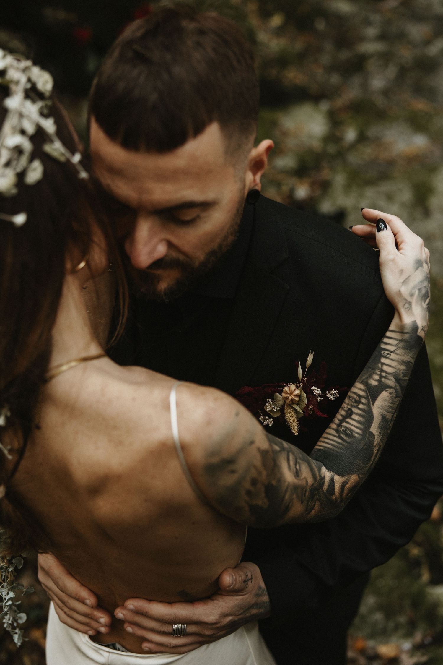 Tattooed groom in black suit embracing bride in backless dress outdoors.