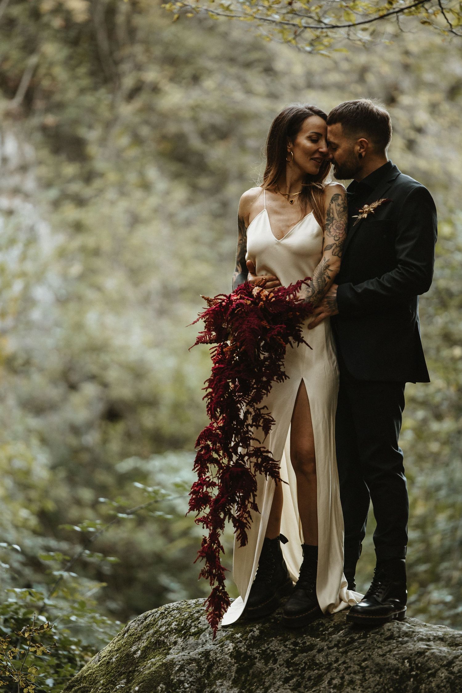 Couple embracing on a rock in forest, bride holding deep red bouquet.