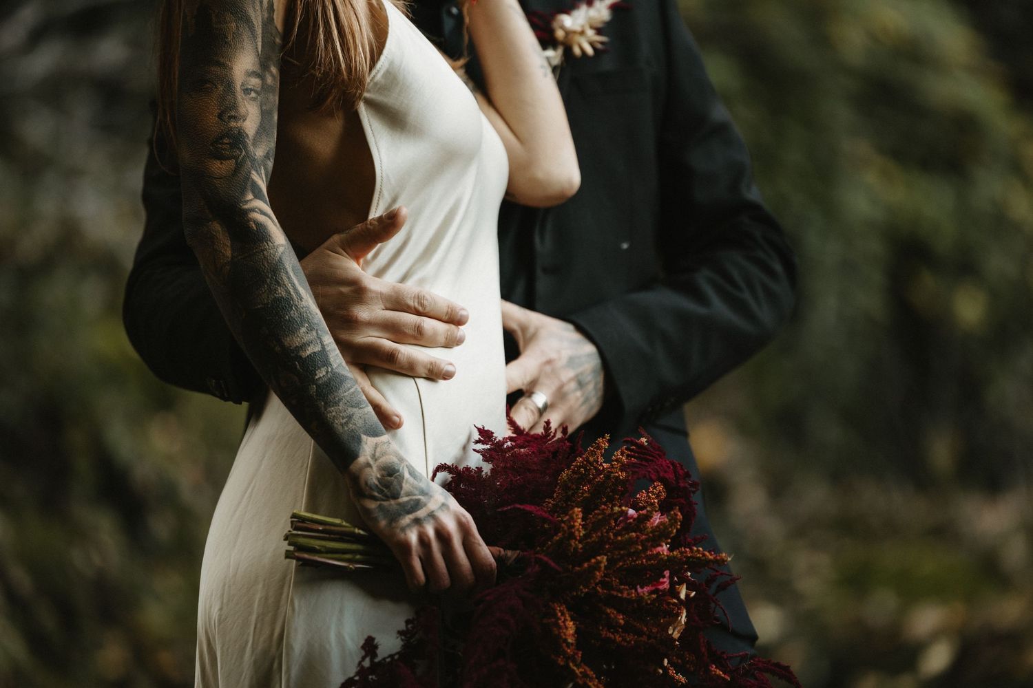 Tattooed couple embracing with bride in white dress holding red bouquet.
