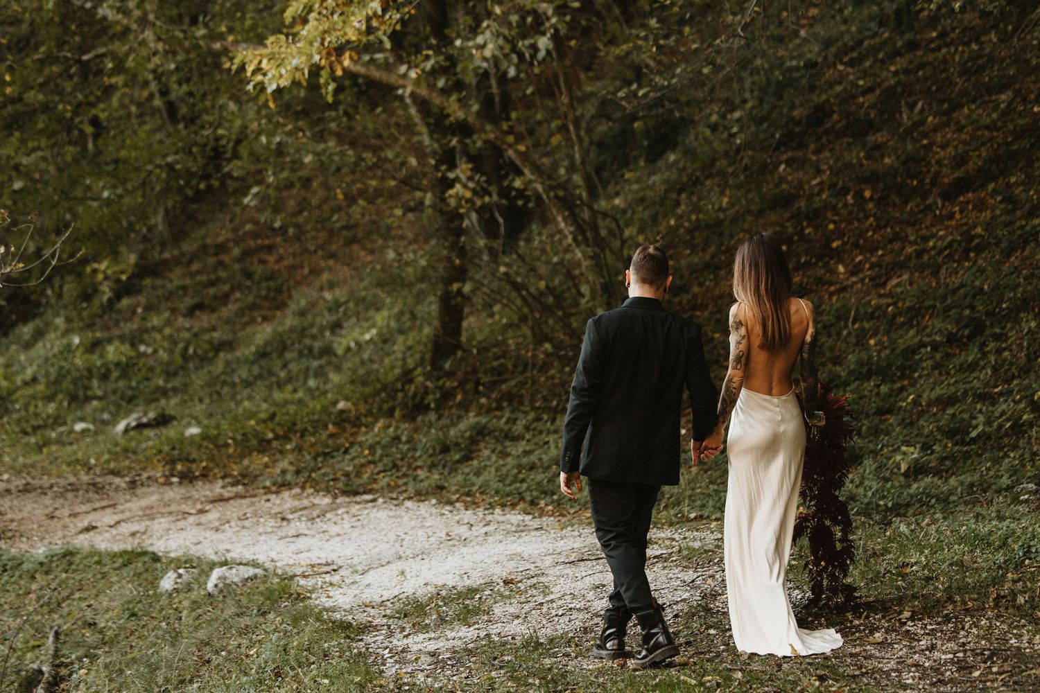 Bride and groom walking hand in hand through a green forest path.