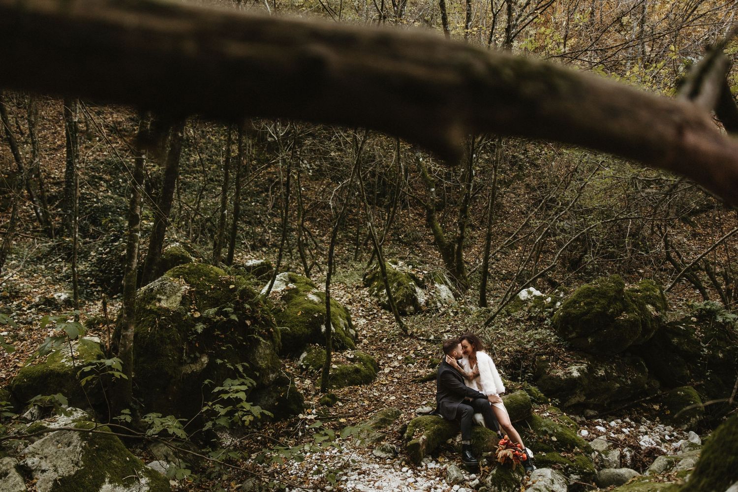 Couple embracing on mossy rocks in an autumn forest during an outdoor engagement photo session.