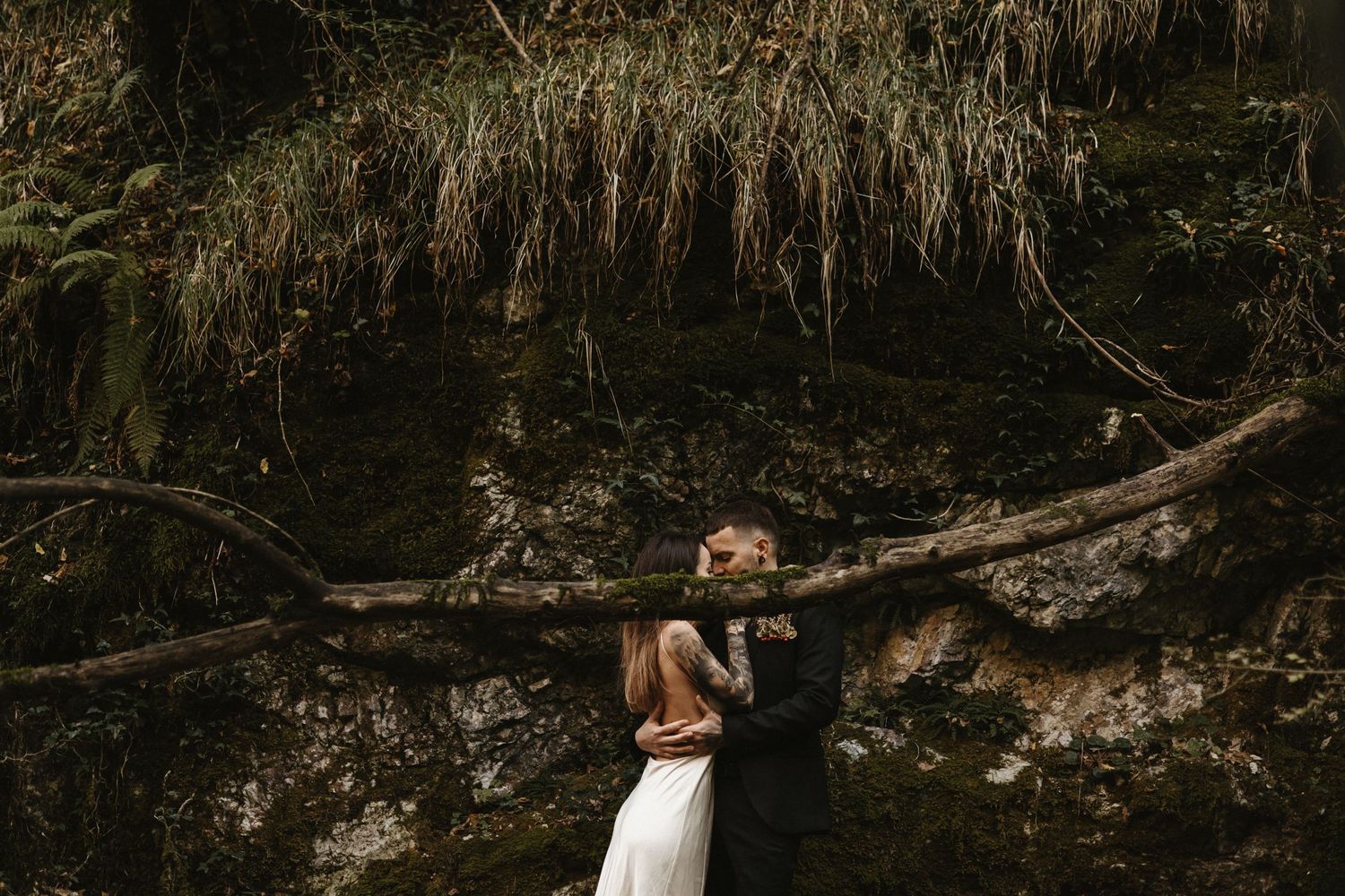 Bride and groom sharing a romantic kiss beside a fallen log in a lush, dark forest setting.