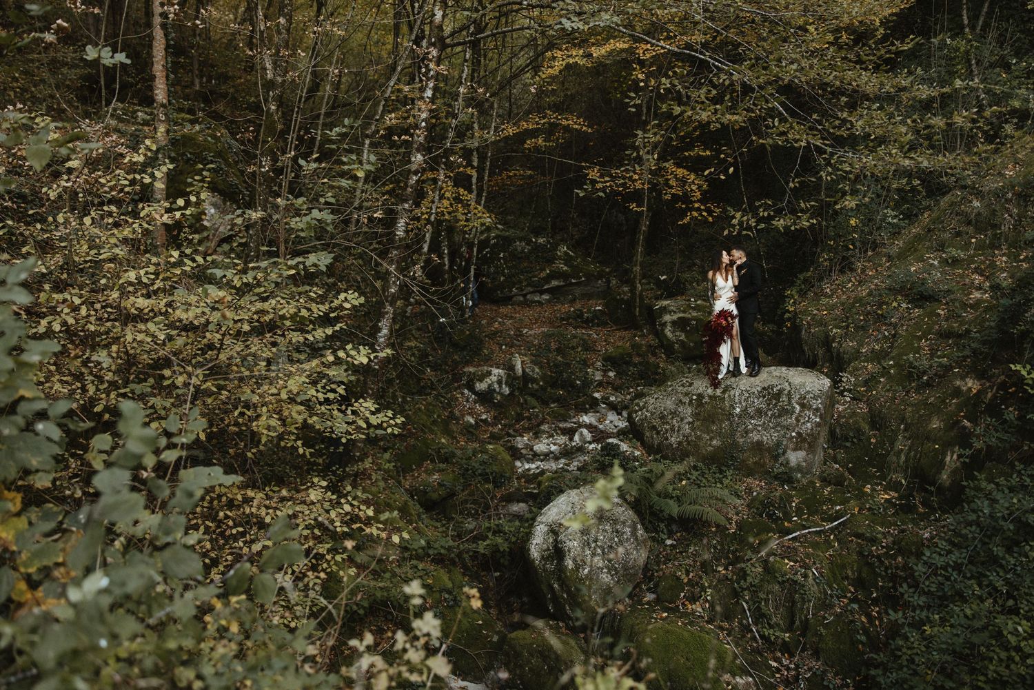Couple standing on boulders surrounded by green foliage near a forest stream in nature.