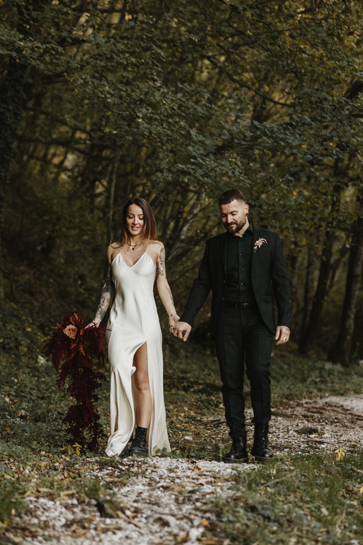 Smiling bride in white slit dress and groom in black suit walking hand-in-hand through a forest.