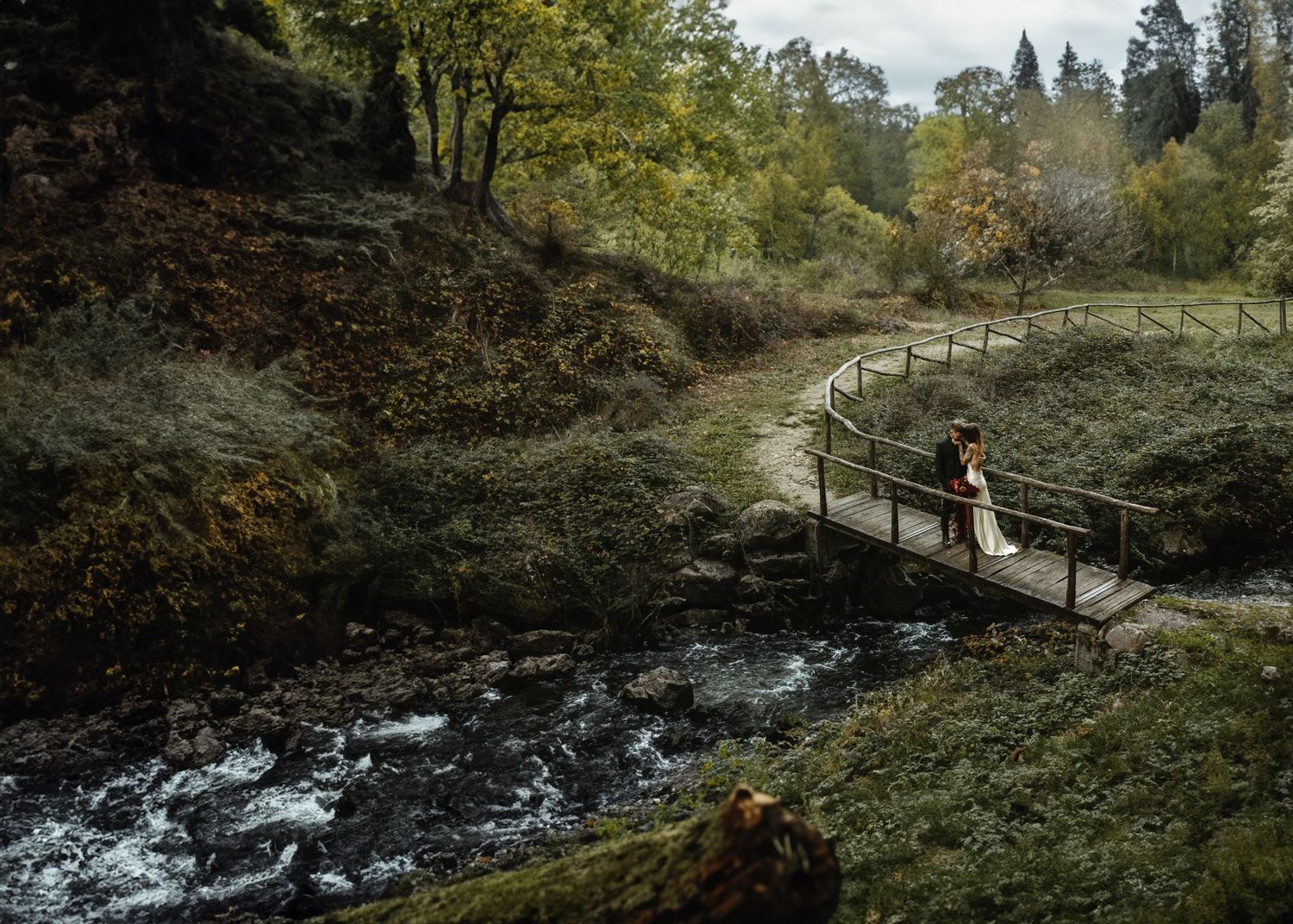 Bride and groom kiss on a wooden bridge over a forest stream in autumn.