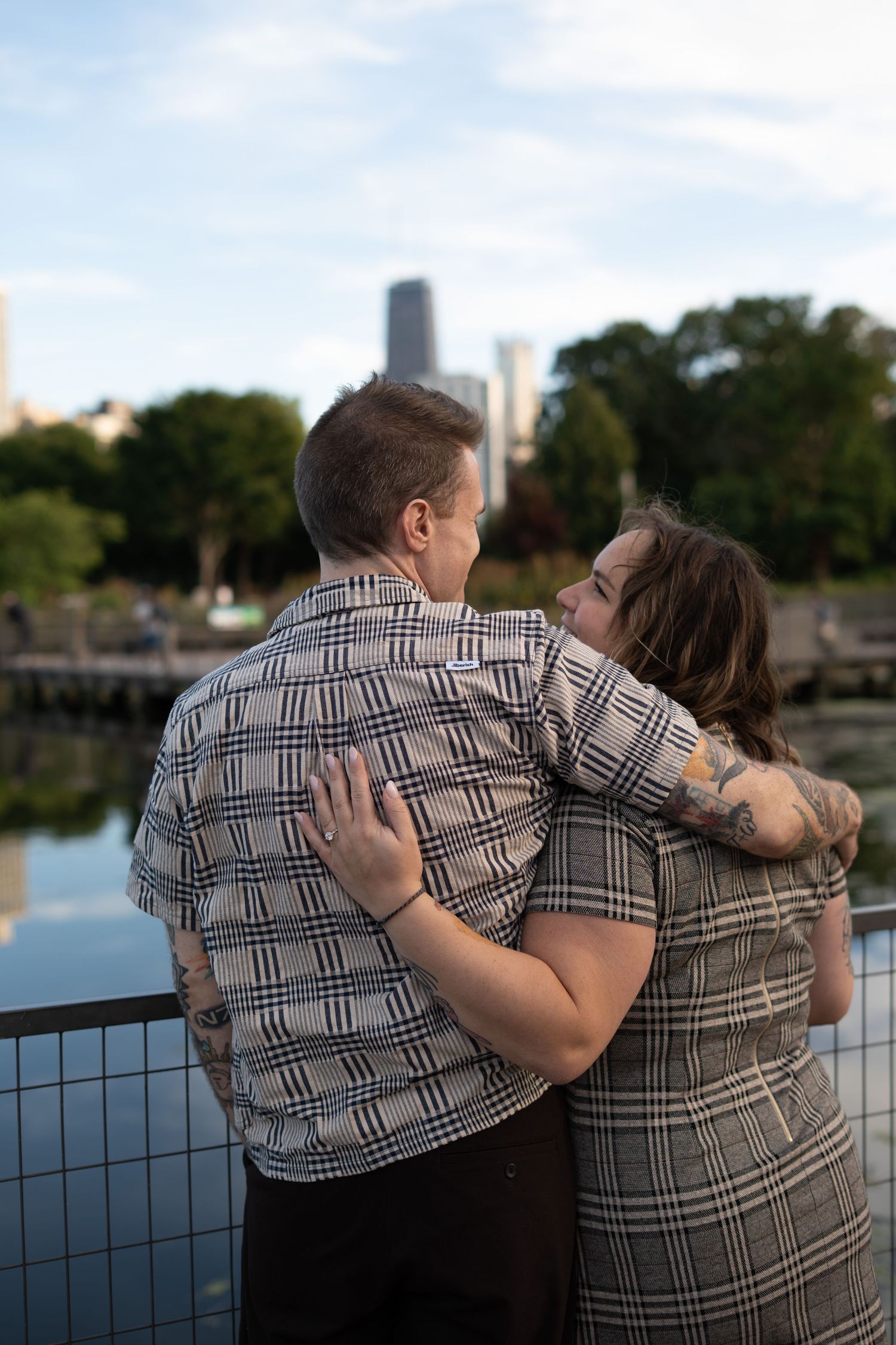 Couple embracing by waterfront with city skyline and trees in background, both wearing plaid outfits.