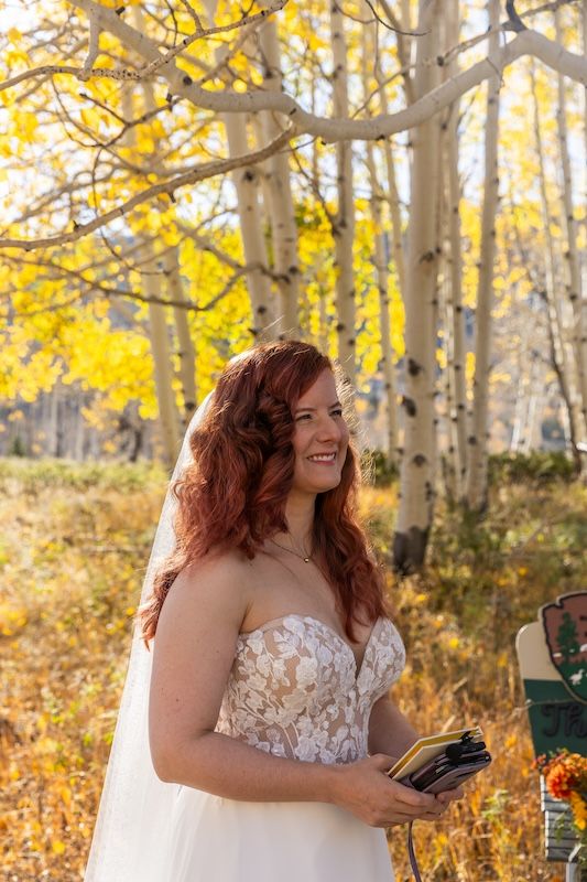 A smiling bride in a lace strapless gown holds vows among golden aspen trees during an autumn outdoor wedding ceremony.