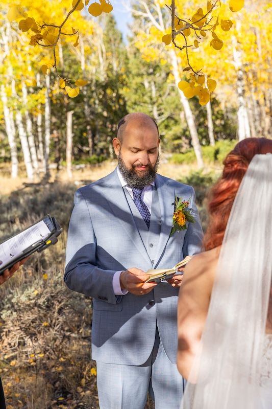 Groom in gray suit reads vows outdoors amid golden aspen trees during fall wedding ceremony.