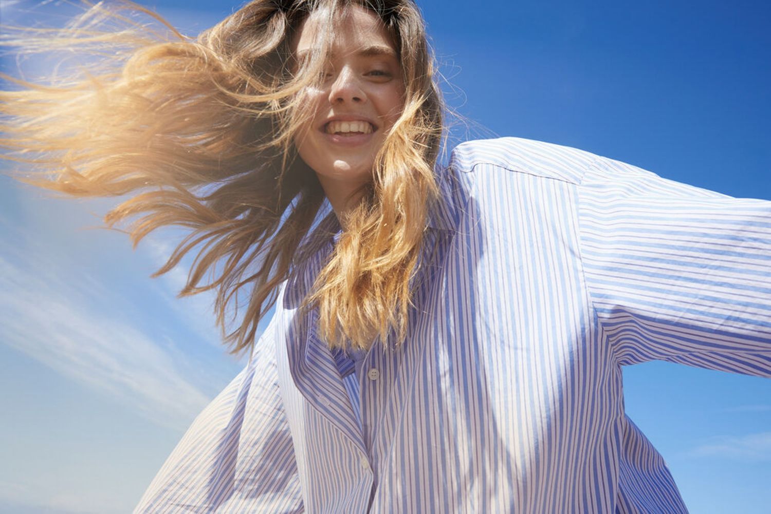 A joyful woman with windswept blonde hair wearing a striped shirt against a bright blue sky.