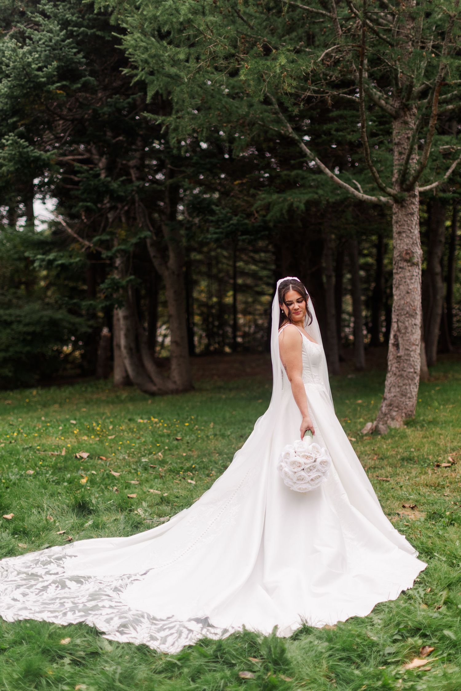 Bride in a white satin wedding gown with a long lace-trimmed train and veil holding a white bouquet on a grassy lawn among evergreen trees.