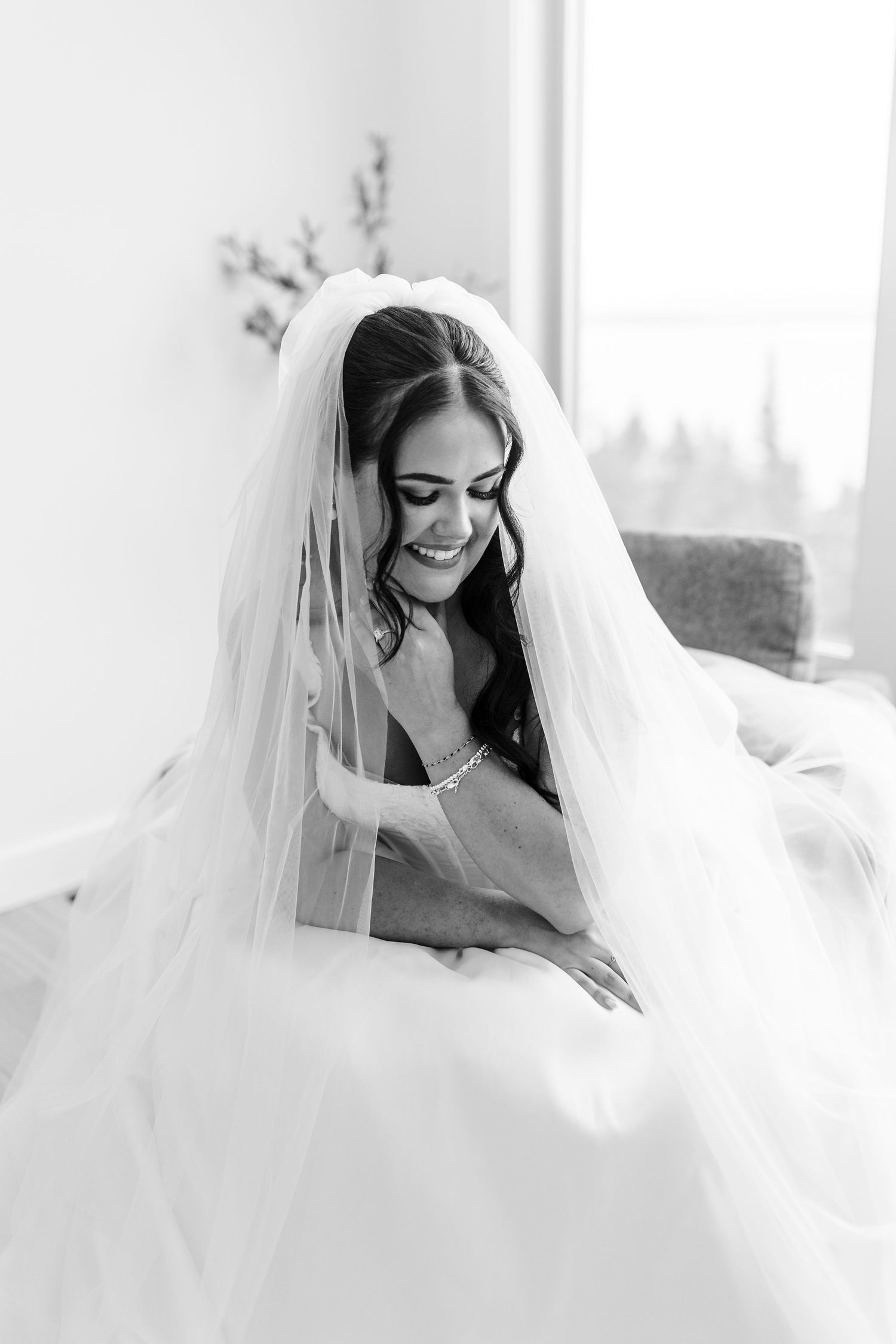 Black and white portrait of a smiling bride in a flowing veil and gown, seated by a bright window with her eyes cast down.