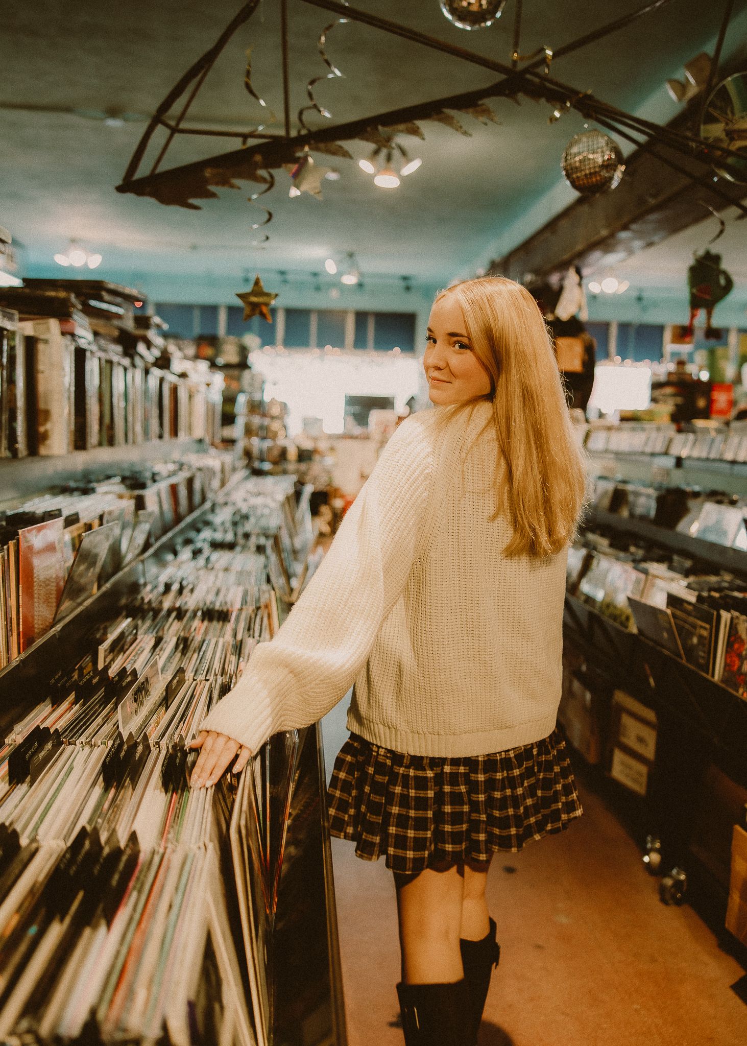 Woman with long blonde hair in a cream sweater and plaid skirt browsing vinyl records in a cozy record store.