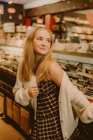Smiling blonde woman in a cream knit cardigan and plaid dress stands beside rows of vinyl records in a cozy music shop.