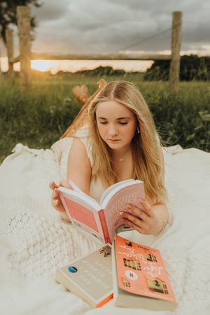 Young woman lying on a white blanket reads a book beside a stack of novels in a field at sunset.