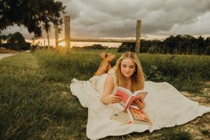Young woman lies on a white blanket in a green field at sunset, reading a pink book beside a wooden fence under dramatic clouds.
