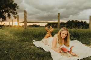 Woman in a white dress and cowboy boots lies on a blanket in a grassy pasture reading a book under a cloudy sunset sky.