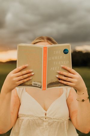 Woman in a cream sundress holds the book The Summer I Turned Pretty over her face beneath a moody sunset sky.