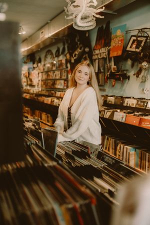 Smiling woman in a white cardigan stands among record bins in a colorful vintage music store filled with posters and decor.
