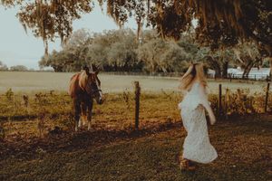 Woman in a flowing white dress walks across a grassy pasture toward a brown horse standing near a wooden fence.