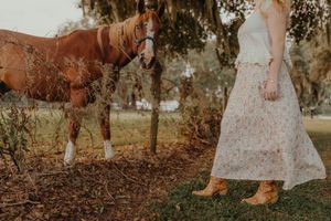 Woman in a floral skirt and tan cowboy boots stands near a chestnut horse beneath a mossy tree in a countryside setting.