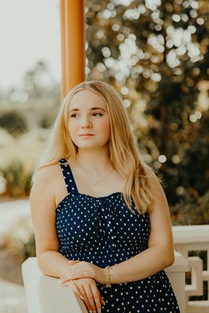 Young woman with long blonde hair in a navy polka dot sundress leans on a white railing, gazing aside against a sunlit, blurred garden.