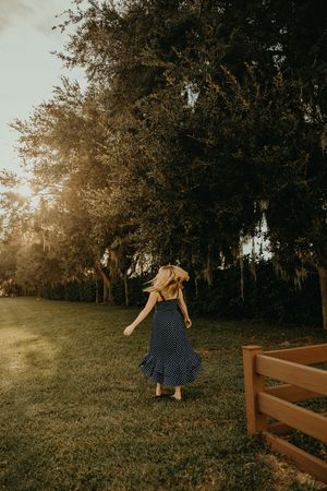Woman in a navy polka dot dress twirls barefoot on a grassy lawn near a wooden fence as sunlight streams through the trees.