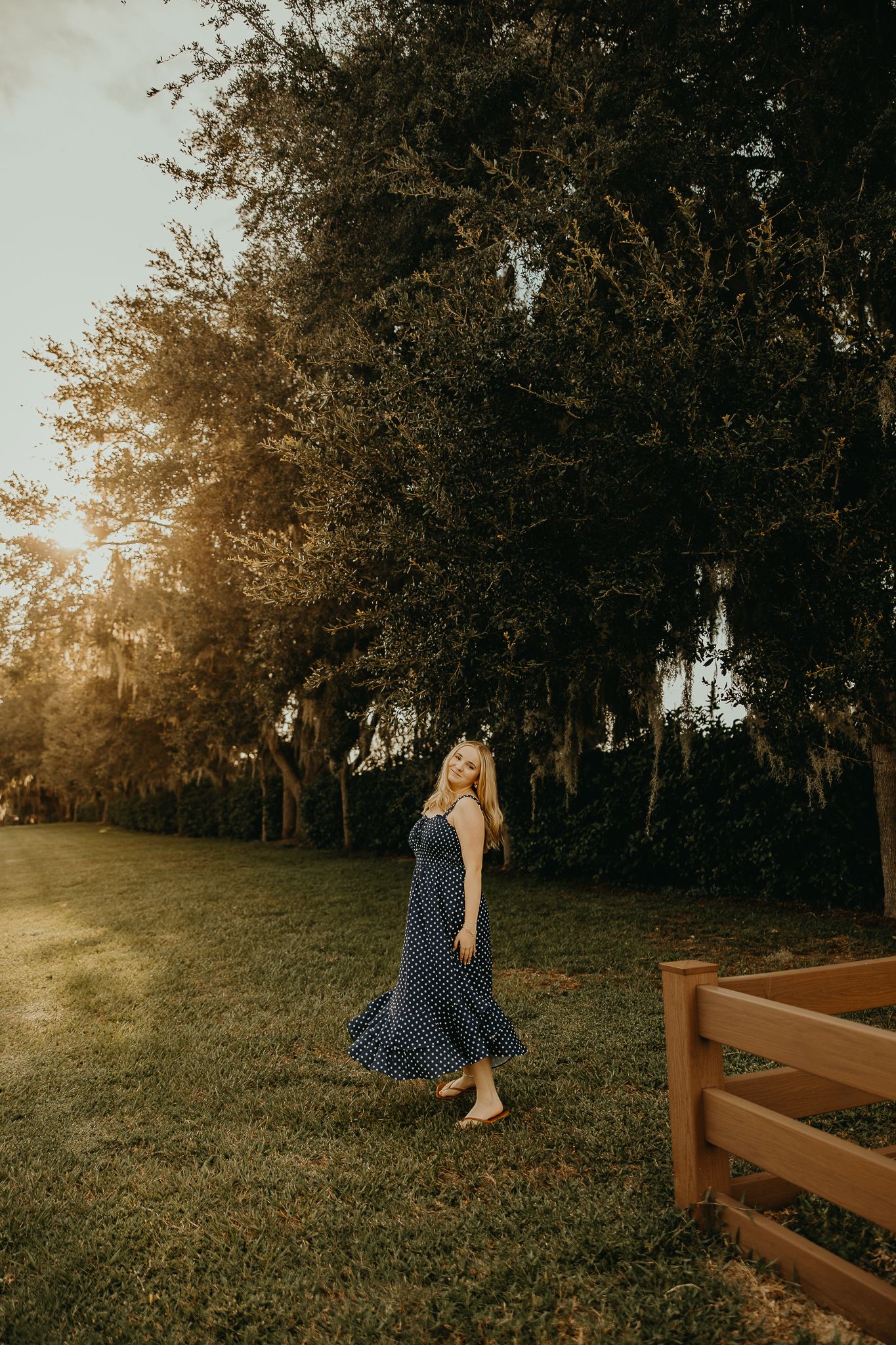 Woman in a navy polka dot maxi dress walks barefoot across a grassy lawn lined with oak trees in warm golden hour sunlight.