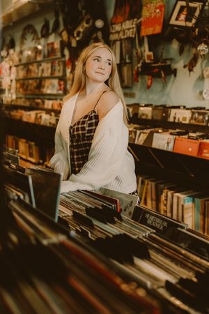 Blonde woman in a white cardigan and plaid top browses crates of vinyl records in a vintage record store with posters on the wall.