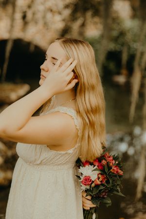 Woman with long blonde hair in a white sundress holds a bouquet of pink and white flowers while touching her hair outdoors.