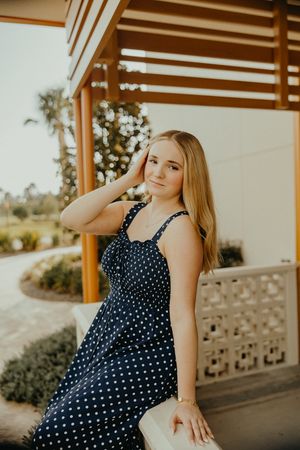 Young woman in a navy polka dot sundress leans on a white ledge under a wooden pergola, hand in her hair.