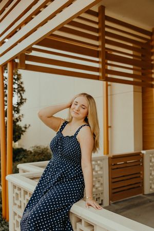Smiling woman in a navy polka dot sundress rests her hand in her hair while sitting on a white ledge beside a wooden pergola.