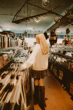 Woman in a cream sweater, plaid skirt and tall brown boots browses vinyl record bins in a record store with hanging disco balls.