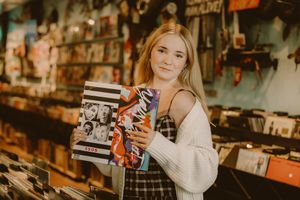 Smiling young woman in a white cardigan and plaid dress holds vinyl records while browsing a colorful record store.