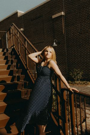 Young woman in a navy polka dot dress poses with her hand in her hair on a sunlit brick staircase with metal railings.