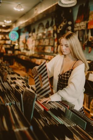 Blonde woman in a white cardigan and plaid top browsing vinyl records in a cozy record store with shelves of albums behind her.