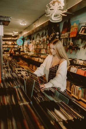 Blonde woman in a white cardigan and plaid dress browses vinyl records in a cozy record store with posters and shelves.