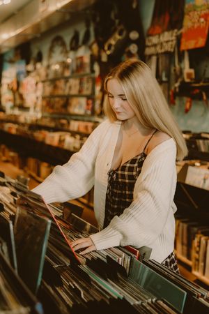 Blonde woman in a white cardigan and plaid dress browses vinyl records in a cozy record store with shelves of albums and posters.