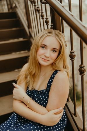 Smiling young woman in a navy polka dot dress sits on outdoor wooden stairs with her arms crossed beside a metal railing.