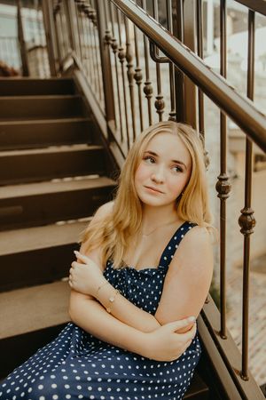 Blonde girl in a navy polka dot dress sits on outdoor stairs with arms crossed beside a metal railing in soft natural light.