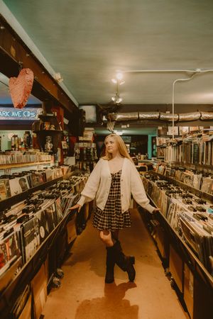 Woman in a white cardigan, plaid dress and black boots standing in the aisle of a vintage vinyl record store lined with shelves.