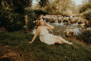 Young woman in a cream dress sits on green grass beside a rocky stream with small waterfalls in a sunlit park.