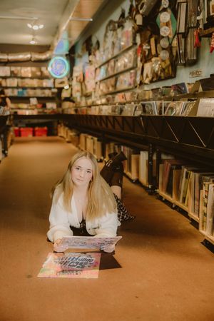 Young woman with long blonde hair lies on the floor of a vinyl record store, holding an album among shelves of records.