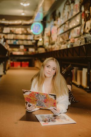 Young woman with long blonde hair lies on the floor of a record store holding a colorful vinyl album cover beside another record.