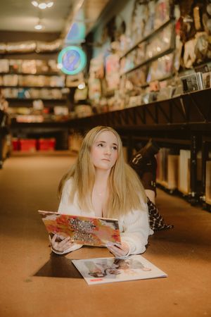 Blonde woman lying on the floor of a record shop holding a colorful vinyl album, with another record on the ground beside her.