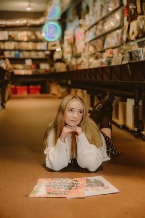 Young girl with long blonde hair lies on the floor of a record store, chin resting on her hands beside two vinyl albums, shelves behind her.