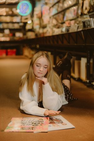 Young woman with long blonde hair in a white knit sweater lies on the floor of a record store, gazing down at vinyl albums.
