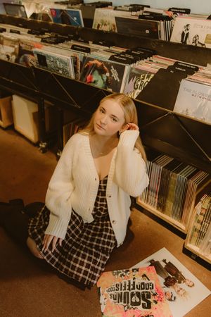 Young woman in a white cardigan and plaid dress sits on the floor of a vinyl record store, surrounded by shelves of albums.