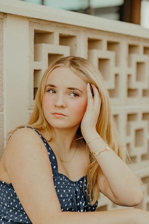 Blonde teenage girl in a navy polka dot dress rests her head on her hand beside a decorative geometric concrete block wall.