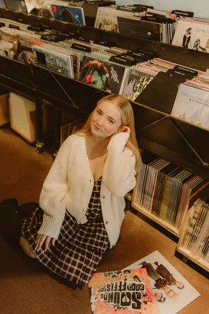 Smiling woman in a white cardigan and plaid dress sits on the floor of a record shop beside bins of vinyl albums.